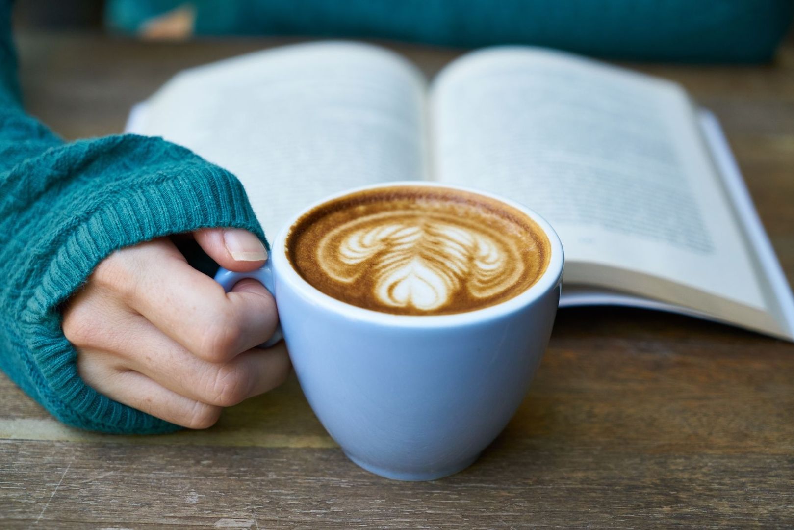 En hand i stickad tröja håller en kaffekopp framför en öppen bok
